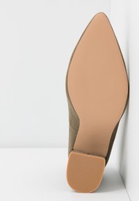 Chaussure à talons hauts en daim vert olive avec un nez pointu et une semelle en caoutchouc beige texturée. Le design du talon présente une silhouette élégante.