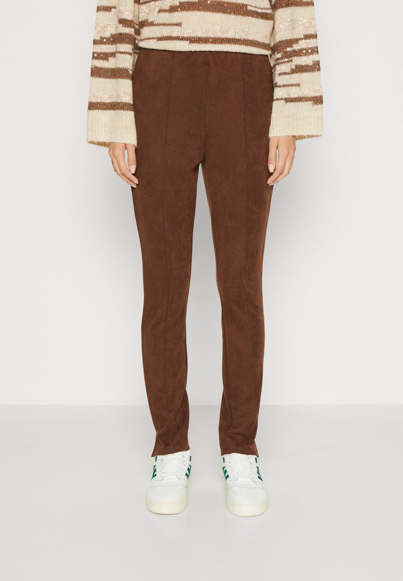Brune suedebukser med en slim fit, som har en glatt tekstur og subtile folder. Kombinert med hvite joggesko med grønne detaljer.
