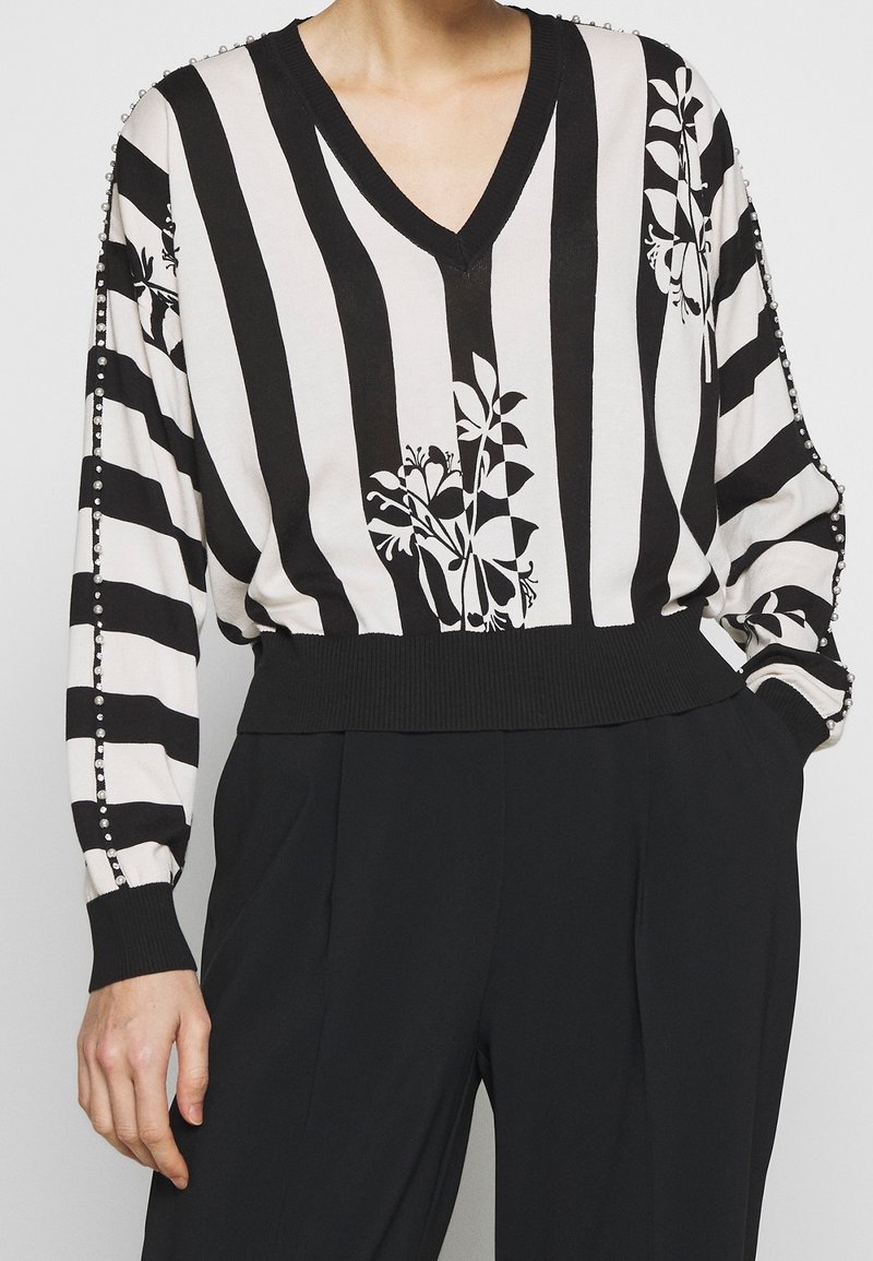 Blouse à col en V à rayures noires et blanches avec des motifs floraux et des détails en perles le long des manches, associée à un pantalon noir taille haute.