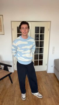 Jonge man die binnenshuis staat, gekleed in een lichtblauwe en witte trui met patroon, donkere broek en witte sneakers, met handen achter de rug.