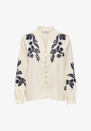 Blouse crème à manches longues avec broderie florale noire sur les épaules et les manches, col et poignets à volants, et boutons recouverts de tissu sur le devant.