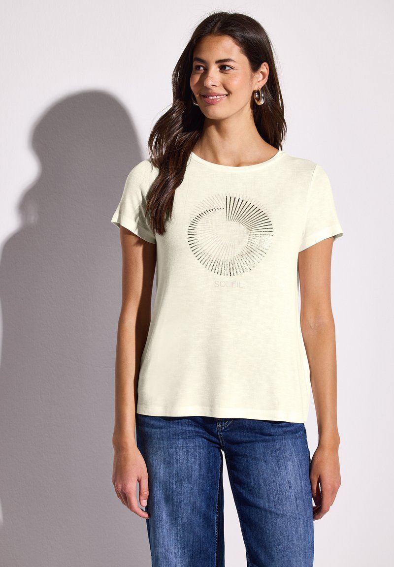 Lys gul kortærmet t-shirt med et cirkulært grafisk design med linjer, der stråler udad, parret med blå jeans. Blød stoftekstur.