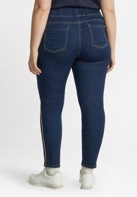 Vaqueros de mezclilla azul oscuro con una cintura alta, dos bolsillos traseros y una franja dorada en el costado. Combinados con zapatillas blancas y una textura de tela visible.