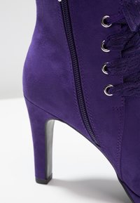 Botín de ante púrpura con tacón alto, que cuenta con una cremallera lateral, parte frontal con cordones y ojales metálicos. Textura suave y forma estructurada.