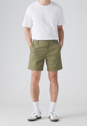 Olijfgroene shorts met een platte voorkant, zijzakken en een iets opgevouwen zoom, gecombineerd met een witte T-shirt met korte mouwen en sneakers.