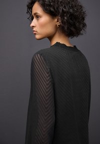 Vestido negro con tejido texturizado y mangas transparentes con patrón de zigzag. Detalle de escote con volantes que realza el diseño. Vista trasera desde un lateral.