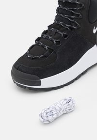 Zwarte suède en mesh sneaker met een witte tussenzool, voorzien van getextureerde veters en een aparte witte en zwarte gevlekte veters.