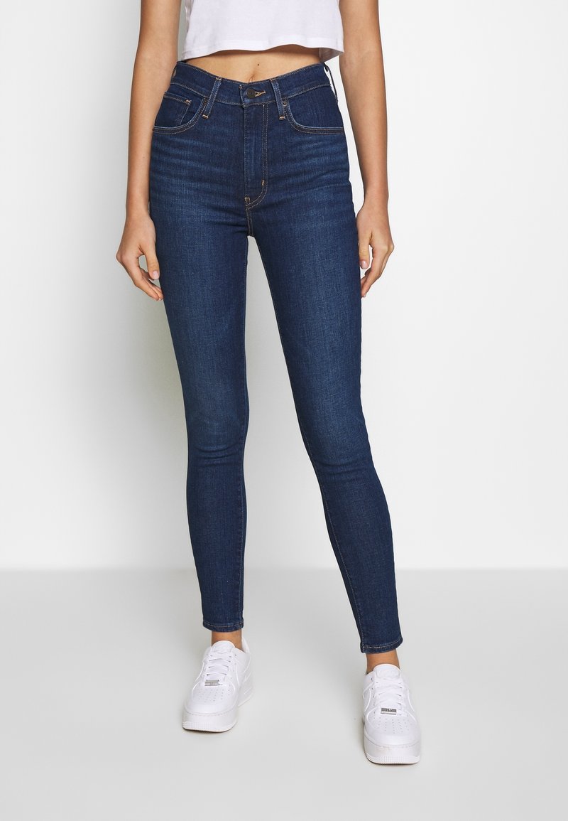 Femme portant un jean skinny bleu foncé taille haute, un crop top blanc et des baskets blanches, debout devant un fond clair uni.