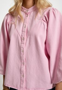 Růžová košile na knoflíky vyrobená z texturované látky. Má vysoký límec, široké rukávy a jemné nařasení na ramenou.