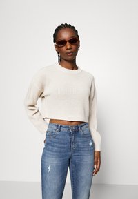 Cremefarvet cropped sweater med ribbestruktur, parret med blå slidte jeans. Modellen bærer brune solbriller og har fletninger.