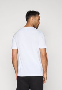 Άνδρας που φοράει ένα απλό λευκό μπλουζάκι κοντομάνικο και μαύρα παντελόνια, στέκεται με την πλάτη στραμμένη και το κεφάλι γυρισμένο στο πλάι, μπροστά από έναν απλό φόντο.