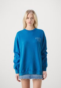 Blauwe katoenen sweatshirt met lange mouwen en een geribbelde ronde hals. Bevat de geborduurde tekst "Tour d'Europe Frankrijk 1995" op de borst.