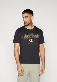 Μαύρο βαμβακερό t-shirt με στυλιζαρισμένο κίτρινο λογότυπο "CHAMPION" στο στήθος. Κοντά μανίκια, στρογγυλή λαιμόκοψη, κανονική εφαρμογή.