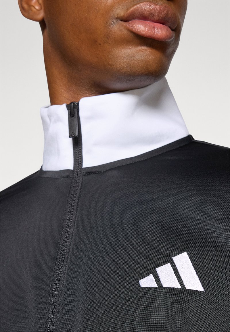 Chaqueta deportiva negra con cremallera y cuello blanco, que presenta un logotipo blanco prominente, confeccionada en una tela suave y texturizada.