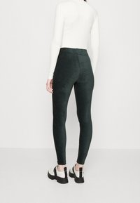 Persona che indossa leggings aderenti verde scuro, top lungo bianco e scarpe chunky bianche, in piedi contro uno sfondo chiaro e uniforme.