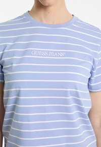 Gaiši zils kokvilnas T-krekls ar baltām horizontālām svītrām. Ar apaļu kakla izgriezumu un izšūtu "GUESS JEANS" logotipu uz krūtīm.