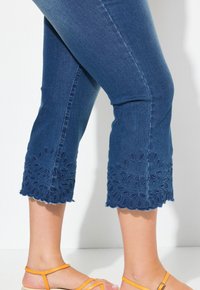 Pantalones recortados de mezclilla en azul oscuro con dobladillos en forma de concha y bordado floral. Diseñados con un ajuste ajustado y bordes deshilachados.