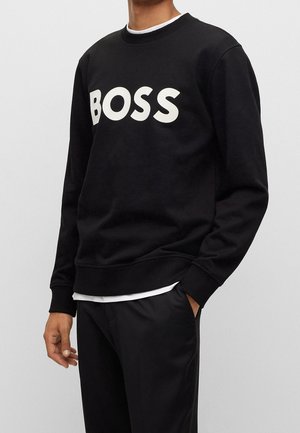Mand iført en sort crewneck-sweatshirt med "BOSS" i store hvide bogstaver på brystet, kombineret med sorte bukser og en hvid undertrøje.