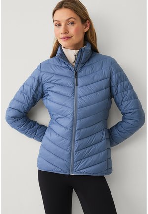 Vrouw met lichtbruin haar, gekleed in een blauw doorgestikt jack, staand met de handen achter haar rug tegen een effen lichte muur.