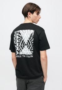 Μαύρο βαμβακερό μπλουζάκι, με μεγάλο λευκό γραφικό στο πίσω μέρος που απεικονίζει αφηρημένα σχήματα και κείμενο που λέει «Αγκαλιάστε το Παράξενο.»