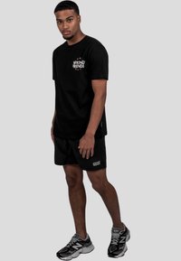 Černé bavlněné tričko s logem "WRONG FRIENDS", spárované s černými šortkami. Model má na sobě šedé a černé sportovní boty. Jednoduchý design, neformální vzhled.