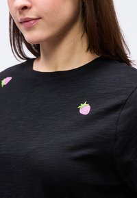 Ženska, oblečena v črno majico z majhnimi vejicami rožnate jagode, izvezene ob ramenih, kjer so vidni njen vrat in spodnji del obraza.