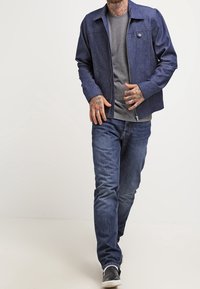 Giacca di denim blu scuro con colletto, zip frontale e tasca sul petto, indossata sopra una maglietta grigia e abbinata a jeans blu e sneakers nere.