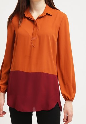 Woman wearing a long-sleeve, two-tone blouse with orange top half, burgundy bottom half, and black pants, standing against a plain background.