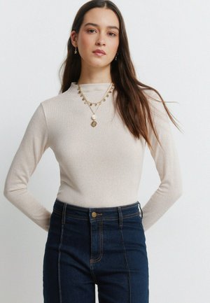 Beiges Langarmshirt mit hohem Hals, kombiniert mit hoch taillierter dunkelblauer Jeans. Dekorativer Goldschmuck und Ohrringe sind zu sehen.