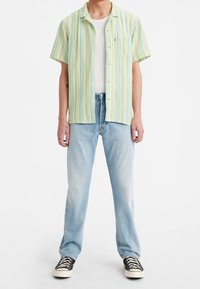 Camisa de manga corta con botones en rayas verdes y amarillas. Se usa sobre una camiseta sin mangas blanca, combinada con vaqueros azul claro y zapatillas negras.