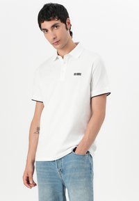 Witte polo met een drieknopen-sluiting, zwarte accenten op de mouwen en een klein zwart logo-detail op de borst. Draagt met lichtblauwe jeans.
