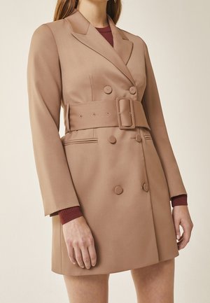 Femme portant une robe blazer ceinturée à double boutonnage couleur beige sur un pull côtelé bordeaux à manches longues, debout devant un fond neutre.