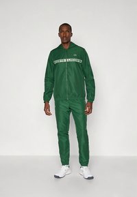 Πράσινη φόρμα από απαλό ύφασμα, που περιλαμβάνει ένα μπουφάν με φερμουάρ και λωρίδα με λογότυπο, καθώς και ελαστικά μανίκια, συνδυασμένη με αντίστοιχο στυλιζαρισμένο παντελόνι.