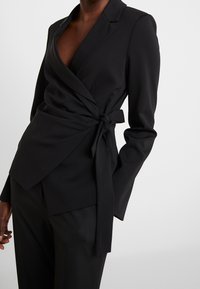 Svart kavaj med långa ärmar, djup V-halsringning och knytning vid midjan; slätt tyg med minimalistisk design och inga synliga mönster.