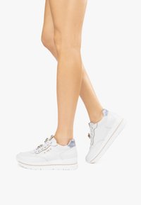 Baskets blanches avec une tige en cuir lisse, accents métalliques gris, laçage plat et une semelle rembourrée avec une bande beige clair.