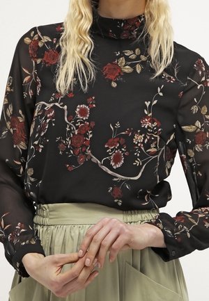 Zwarte doorschijnende blouse met rode bloemenprint en hoge hals, gecombineerd met een licht olijfgroene rok met hoge taille, handen zachtjes in elkaar gevouwen op de taille.