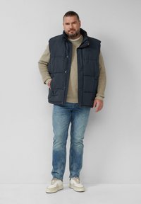 Man met een baard in een marineblauwe puffer vest, een beige trui, blauwe spijkerbroek en witte sneakers staat tegen een effen grijze muur.