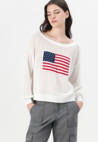 Biely pletený sveter s nadrozmerným strihom, na prednej strane s dizajnom americkej vlajky v červenej a modrej farbe. V páre so šedými cargo nohavicami.