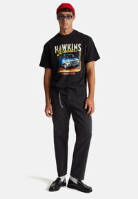 Černé tričko s potiskem "HAWKINS" a barevný motiv vintage dodávky. Kombinováno s černými džínami a černými mokasíny s detailním řetízkem.