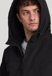 Giacca nera con zip frontale e cappuccio, con fodera testurizzata, zip anteriore e coulisse regolabili. Dettagli della zip argentati visibili.