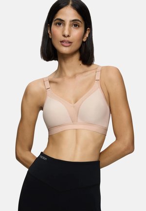 Femme aux cheveux courts et foncés portant un soutien-gorge rose pâle et un pantalon noir taille haute, debout devant un fond clair uni.