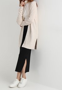 Beige ribbad cardigan med öppen framsida, lagd över en figurnära svart kjol med sidoslits, tillsammans med vita sneakers.
