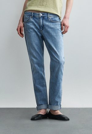 Άτομο που φοράει ανοιχτό μπλε τζιν με γυρισμένα μπατζάκια, μαύρα παπούτσια slip-on και ανοιχτό κίτρινο πουκάμισο, με τα χέρια χαλαρά δίπλα στο σώμα.