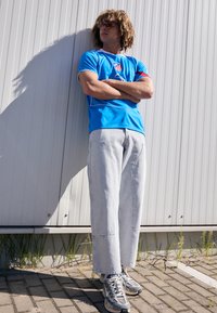 Blue athletic shirt with logo, light gray wide-leg jeans, and gray sneakers with mesh accents, set against a simple background.