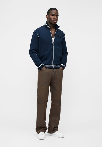 Homme debout les mains dans les poches portant une veste zippée marine, un pantalon marron, des chaussures blanches et un sous-vêtement blanc sur un fond uni.