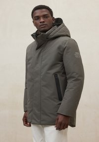 Parka imbottita verde oliva, con cappuccio, tasca con zip nera e marchio discreto sulla manica destra. Tessuto dalla texture liscia.