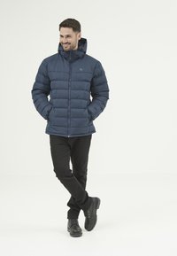 Marinblå pufferjacka med huva, dragkedja och elastiska ärmslut. Bärs över svarta byxor och robusta svarta kängor. Texturerad, isolerad tyg.