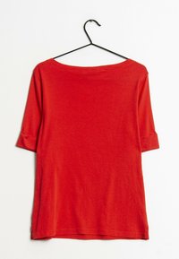 Červené, měkké bavlněné tričko s širokým výstřihem a tříčtvrtečními rukávy, zavěšené na černém háčku. Hladká textura, ležérní střih.
