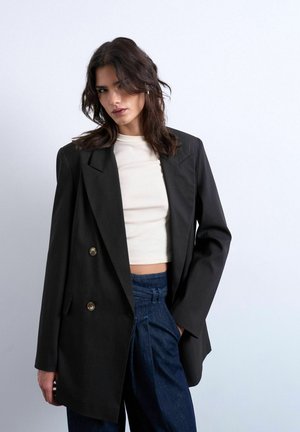 Kvinde med skulderlangt mørkt hår iført en stor sort blazer over en hvid croppet top og højtaljede blå bukser mod en hvid baggrund.