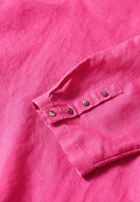 Tessuto di camicia in lino rosa con un risvolto, dotato di cinque bottoni metallici e una superficie liscia, leggermente texture.
